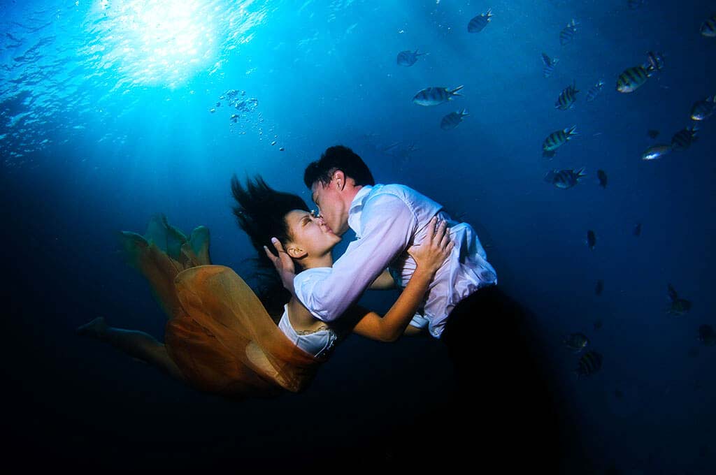 underwater wedding photography for underwater bridal proposal, pre-wedding photography for couples tuckys photography. A couple embracing and sharing a kiss underwater, surrounded by fish, with rays of sunlight penetrating the deep blue sea.
