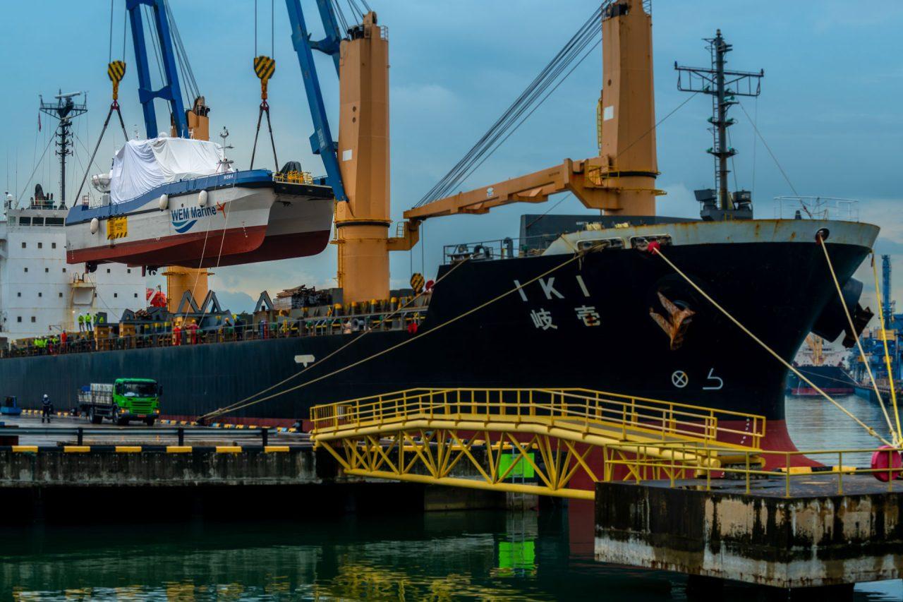 WEM Marine catamaran wrapped on its cradle and lifted by the heavy-lift vessel IKI’s cranes above the wharf, with the yellow gangway and vehicles below