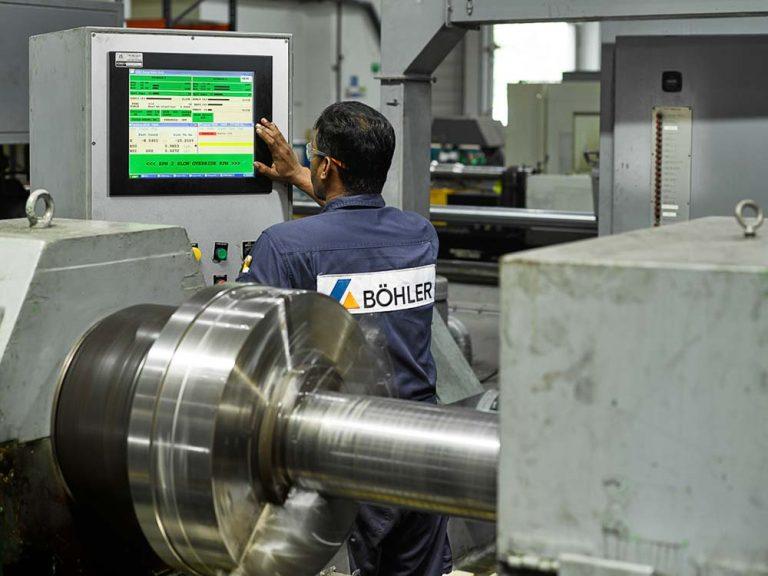A Bohler technician at the CNC control panel with a machined steel shaft on the bed.
