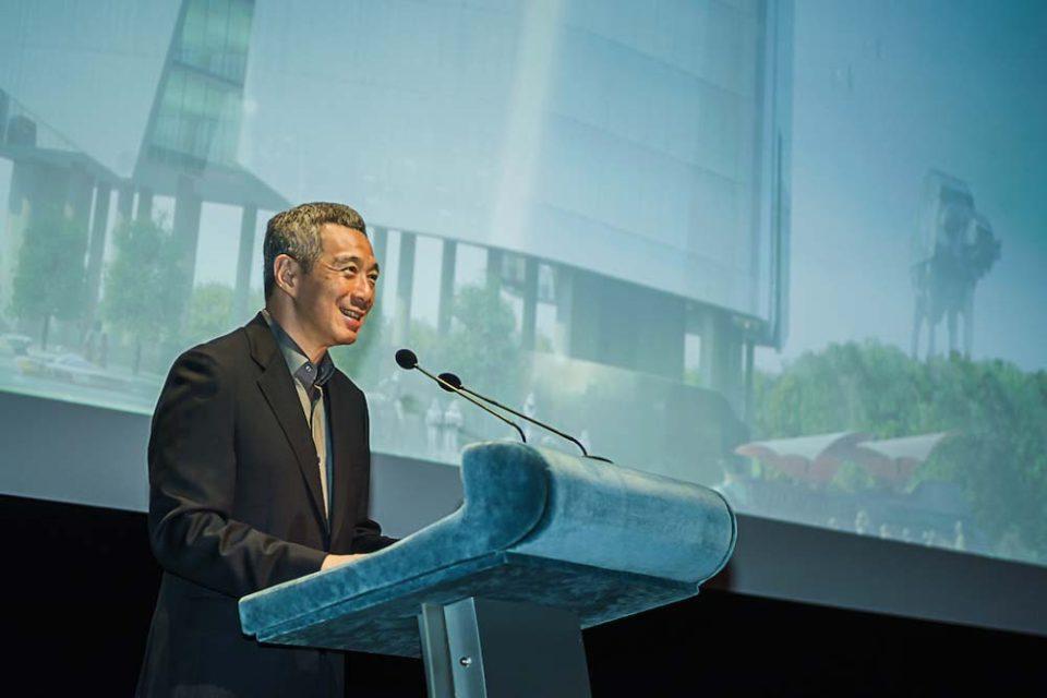 Singapore corporate events photography capturing PM Lee Hsien Loong speaking at a podium with a large projection screen displaying urban architecture and a futuristic structure in the background.