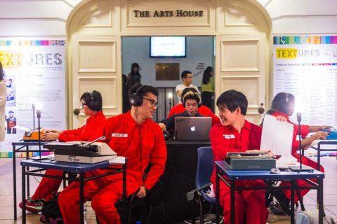 Behind the scene moments during a literature event. Group of individuals in red uniforms working at tables with headphones, laptops, and equipment, set in front of 