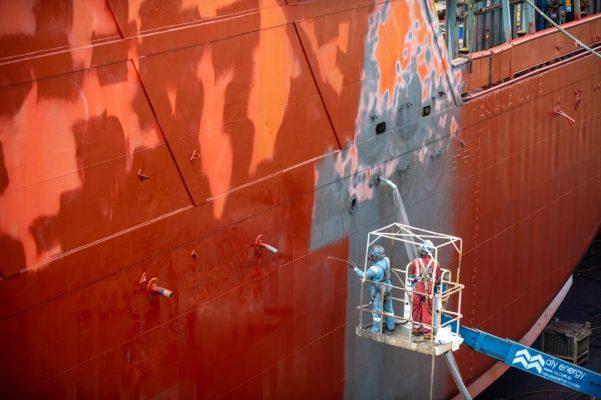 Painters in a boom-lift cradle applying the first protective coat to the blasted hull. Shipyard progress documentation in Singapore.