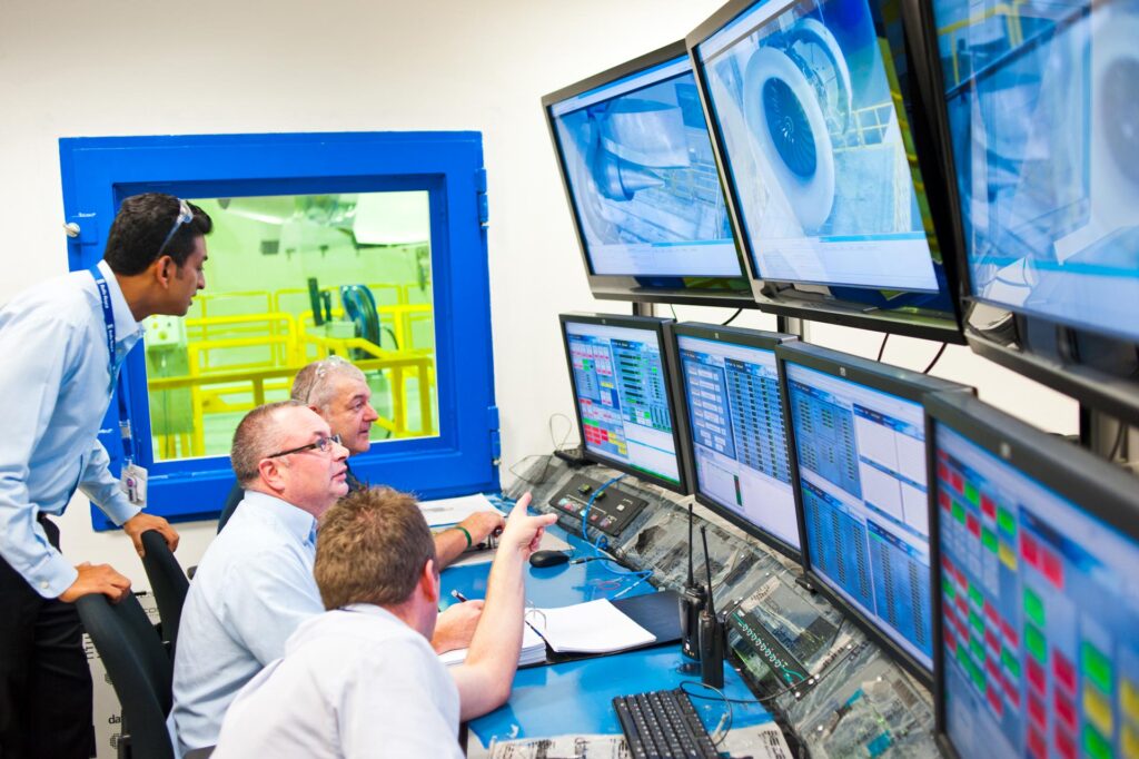 Engineers in the Rolls-Royce Seletar test cell control room watching a bank of monitors, with the engine visible through the blue-framed window