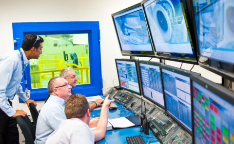 Engineers in the Rolls-Royce Seletar test cell control room watching a bank of monitors, with the engine visible through the blue-framed window