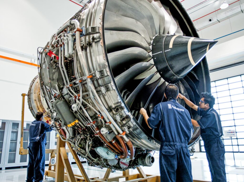 Trent engine on a build stand in the Rolls-Royce Seletar assembly bay, fan and spinner facing right, three technicians working around it