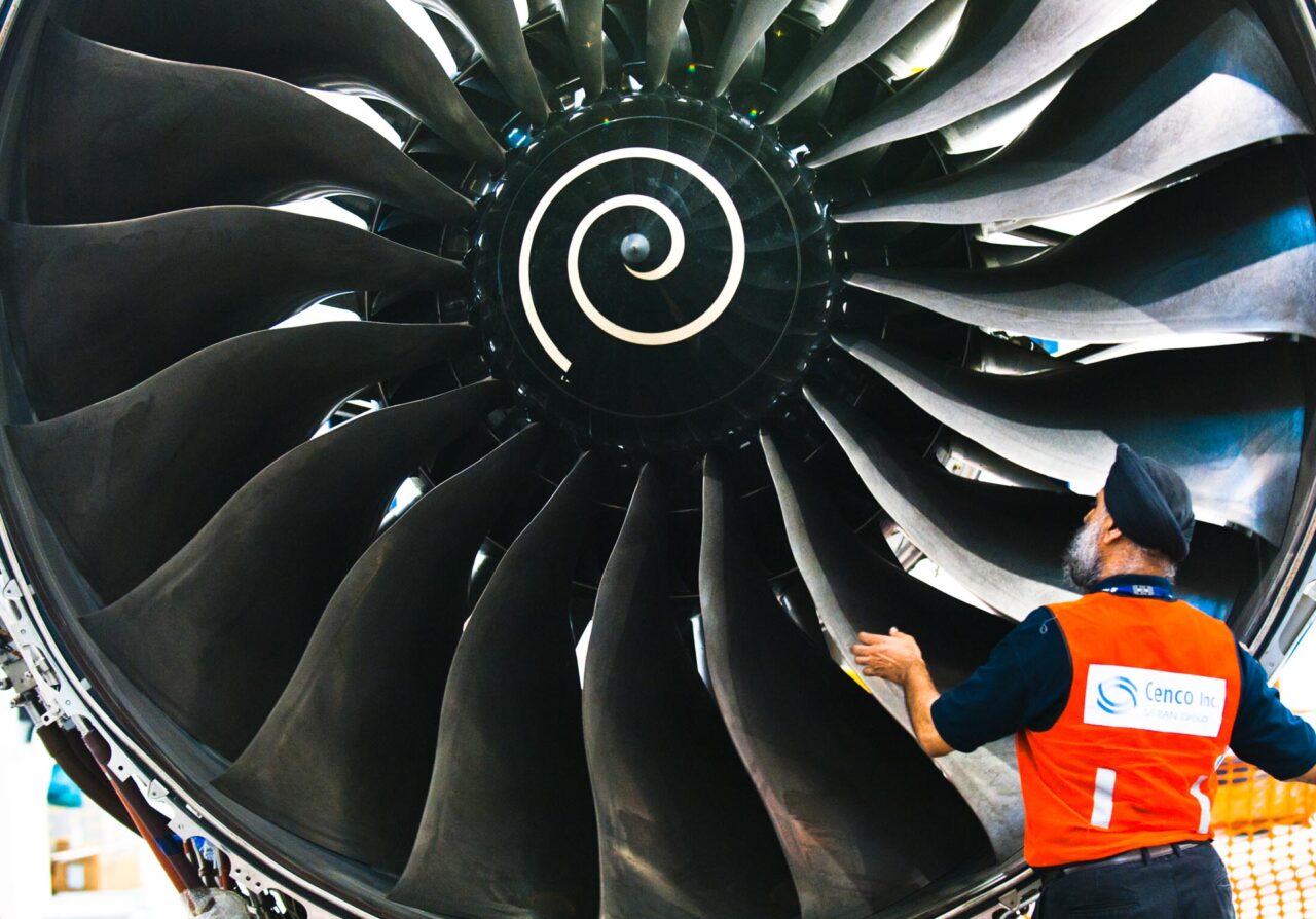 Technician in an orange vest checking the fan blades of a Trent engine by hand, close view of the swirl-marked spinner, Singapore