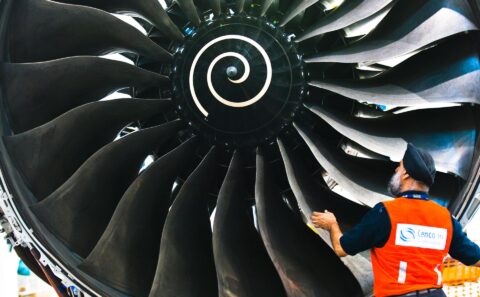 Technician in an orange vest checking the fan blades of a Trent engine by hand, close view of the swirl-marked spinner, Singapore