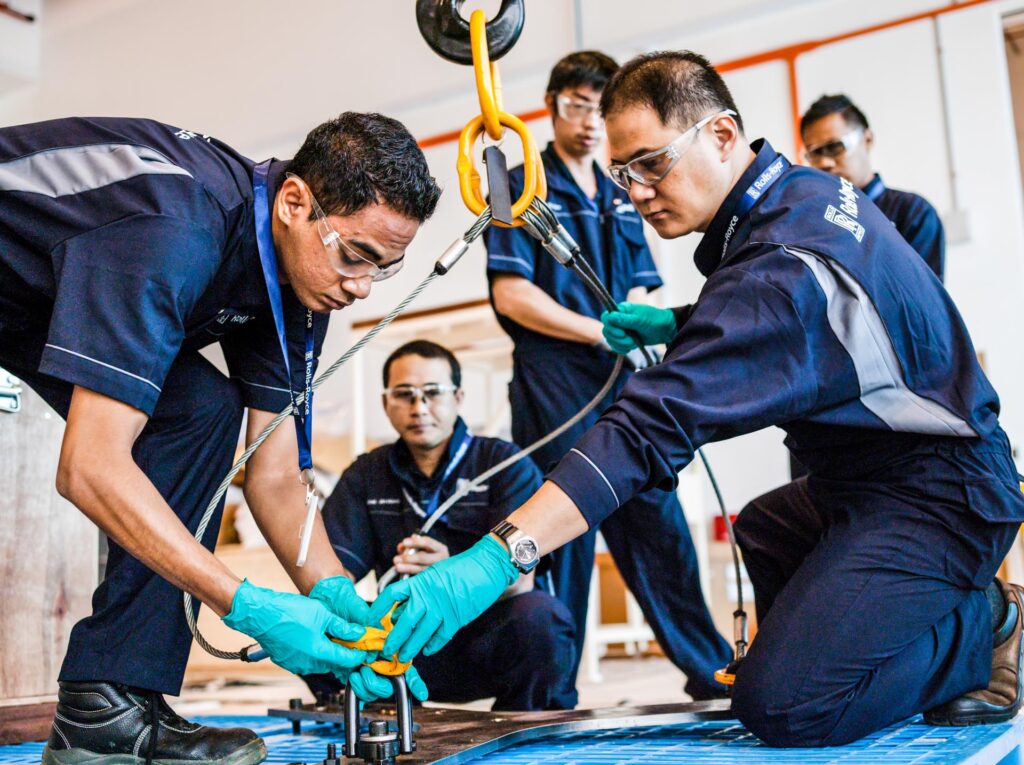 Rolls-Royce trainees in safety glasses and green gloves fitting a lifting shackle to a plate during a rigging class at the Seletar Regional Training Centre, Singapore