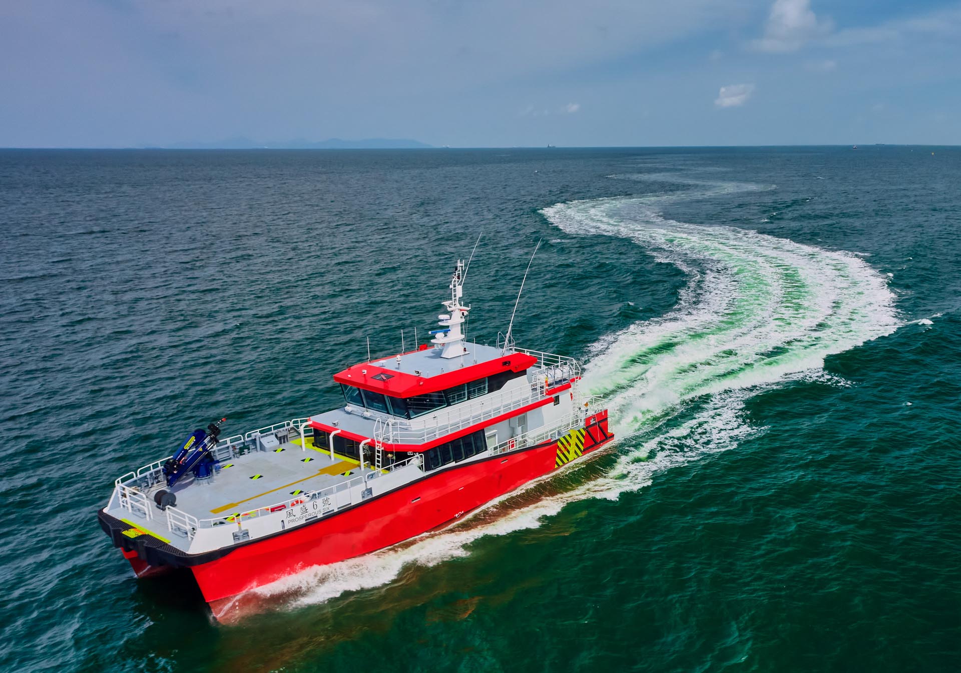 Aerial view of the red and white crew transfer vessel Prosperous 6 turning at speed, its white wake curving across green sea