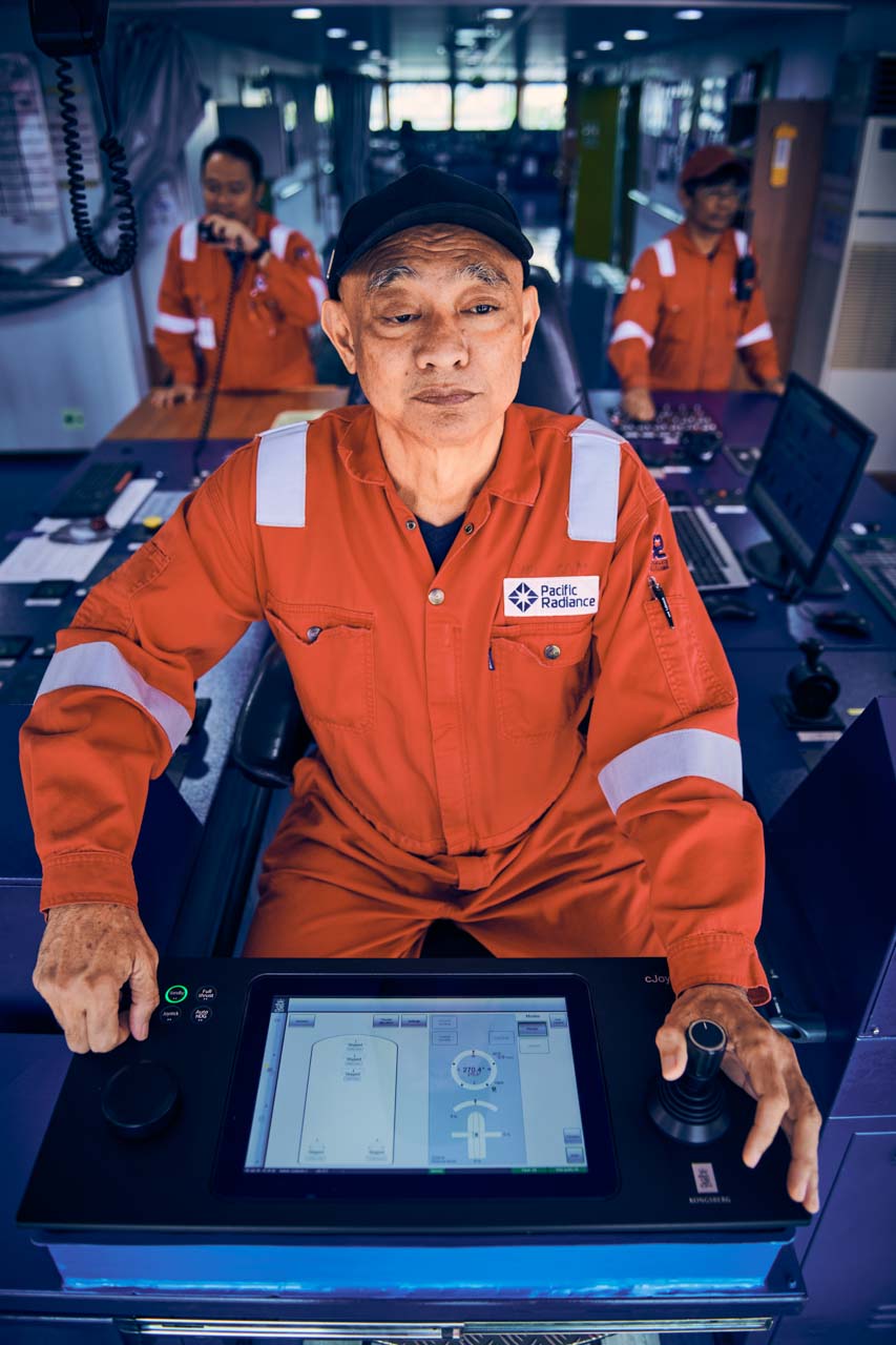 Captain of SAFEEN Centurion 2 in orange Pacific Radiance coveralls at the bridge console, hands on the joystick and control panel, with two crew on the radios behind him