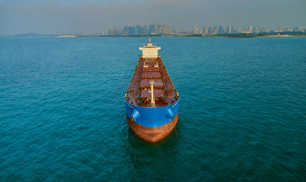 Bulk carrier Safesea Sindoor seen bow-on from the air in Singapore waters, blue bow centred, the city skyline a thin line behind her