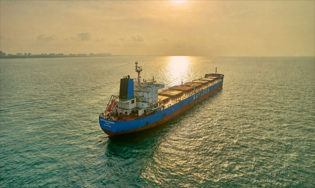 Bulk carrier Safesea Sindoor at anchor off Singapore, backlit at sunset, the sun's reflection running down the water to the hull