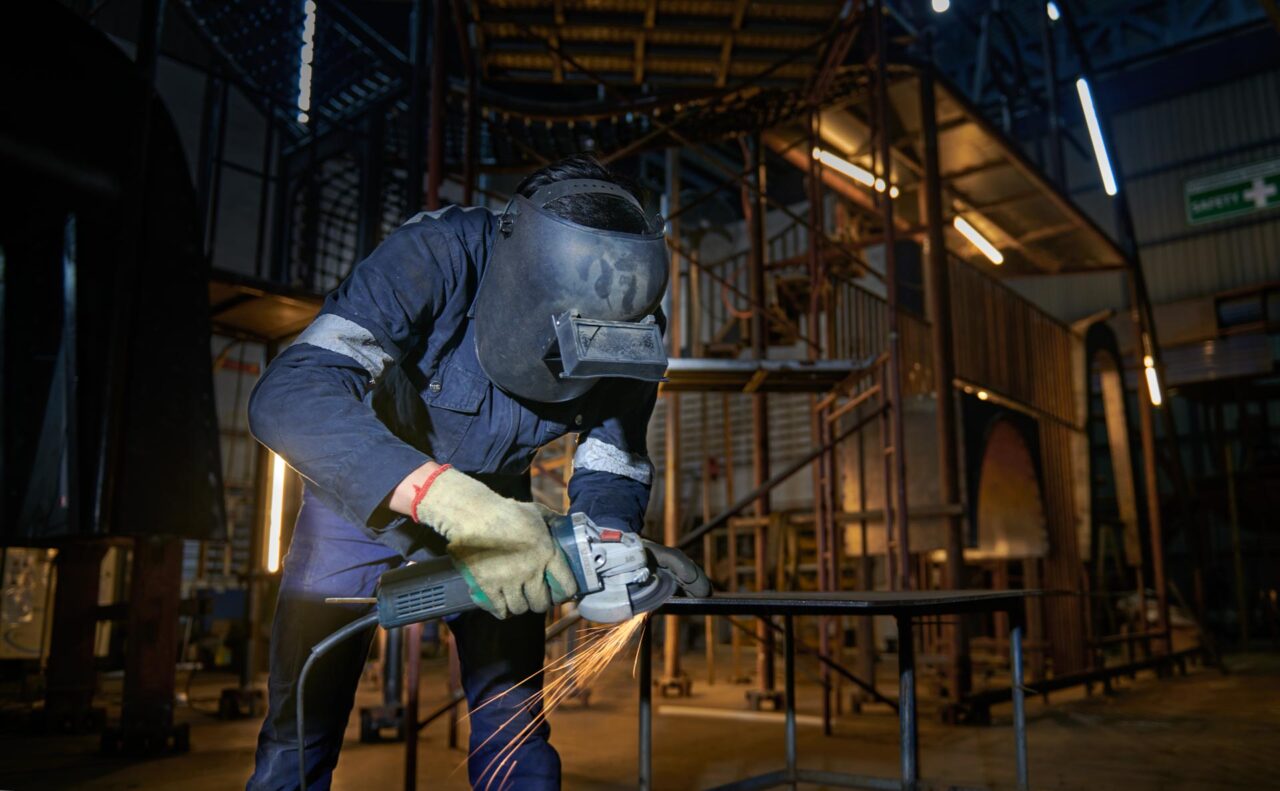 Worker in a face shield and gloves cutting a steel plate with an angle grinder, sparks flying, inside a steel fabrication workshop in Singapore
