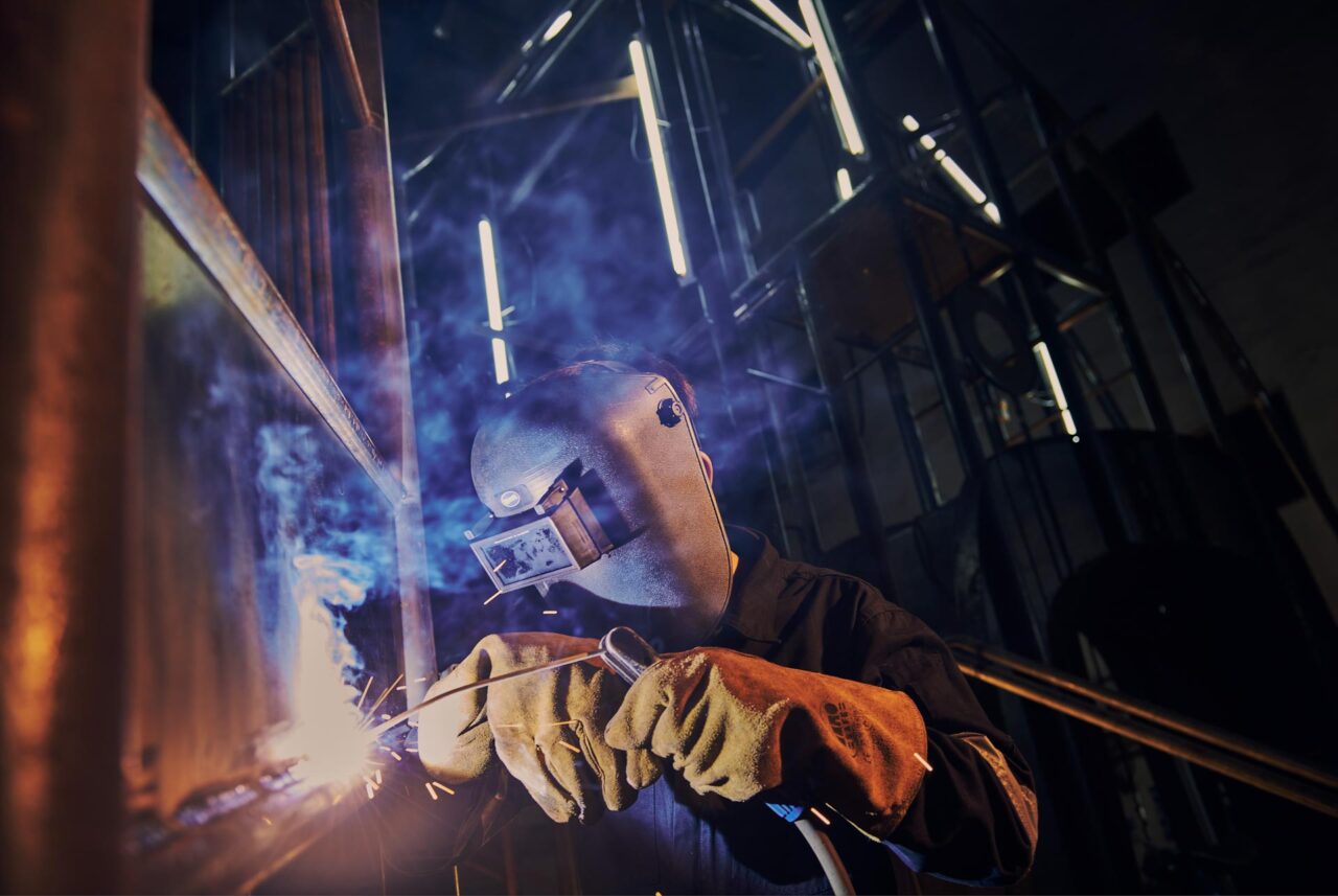 Welder in a flip-up welding mask and leather gloves arc welding a steel frame, blue smoke and sparks, low angle inside a fabrication workshop in Singapore