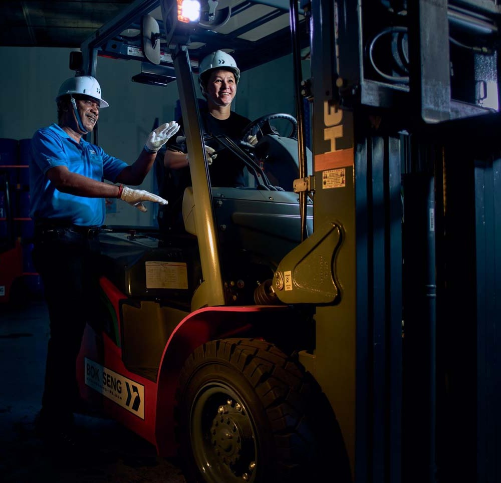 Forklift instructor in a white helmet directing a trainee at the wheel of a red Bok Seng forklift inside a dark warehouse