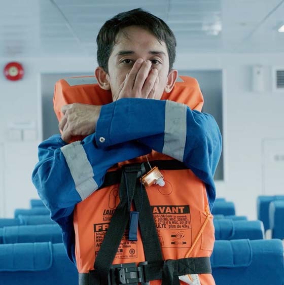 Crew member in a blue boiler suit and orange lifejacket demonstrating the nose-and-mouth hold before an emergency water entry, in the passenger cabin of a vessel