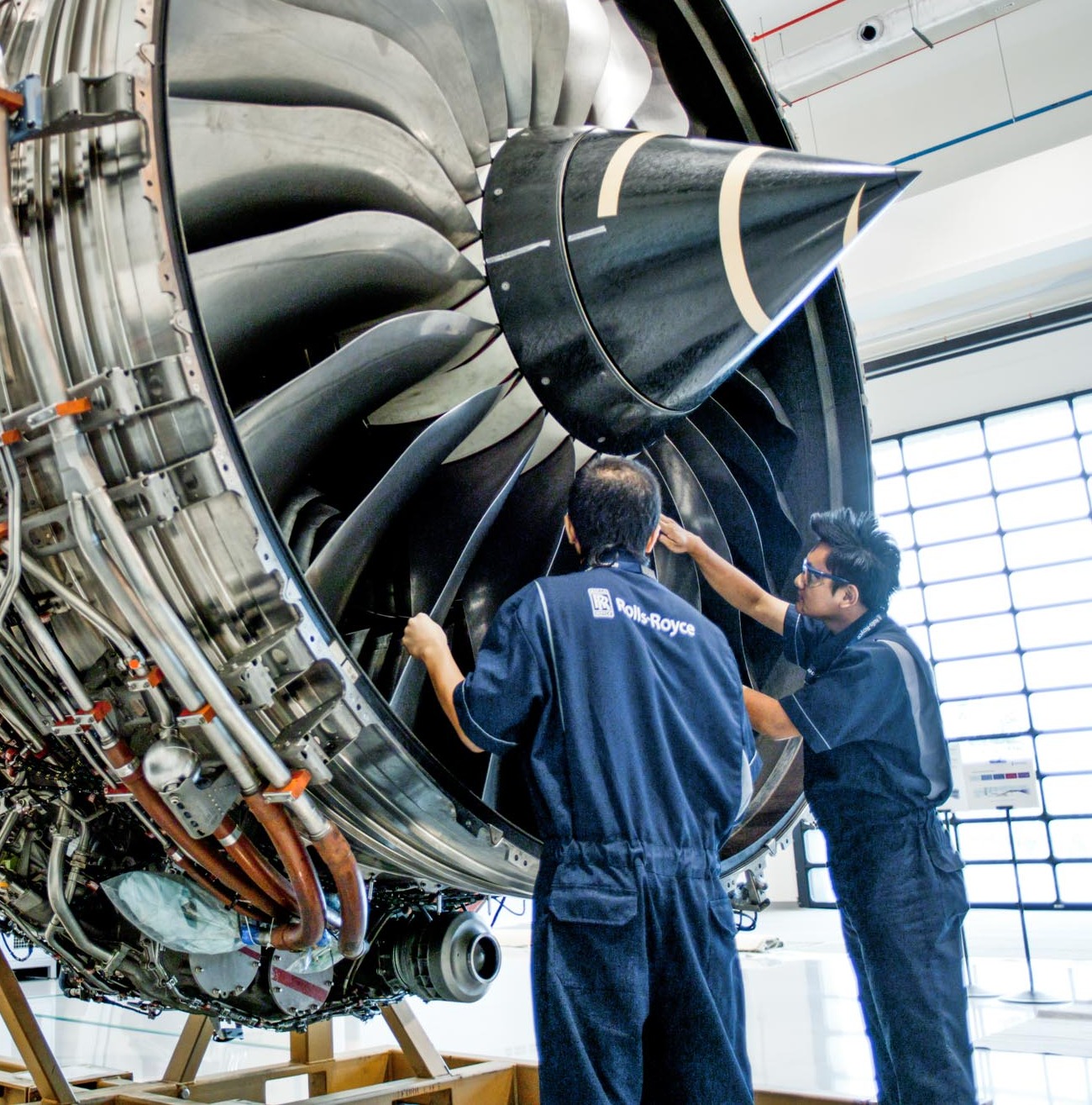 Front of a Rolls-Royce Trent engine on its build stand, spinner and fan blades filling the frame, two technicians in Rolls-Royce overalls working at the blades