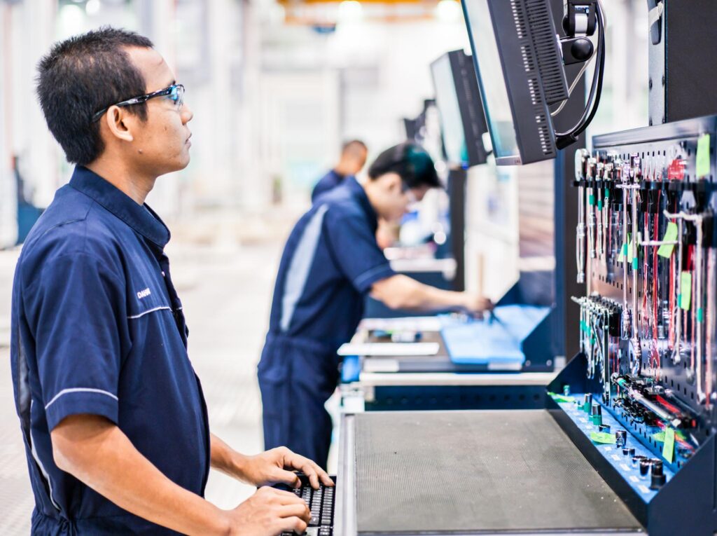 Rolls-Royce technician in safety glasses at an engine assembly workstation with a monitor and a shadow board of tools, Seletar, Singapore