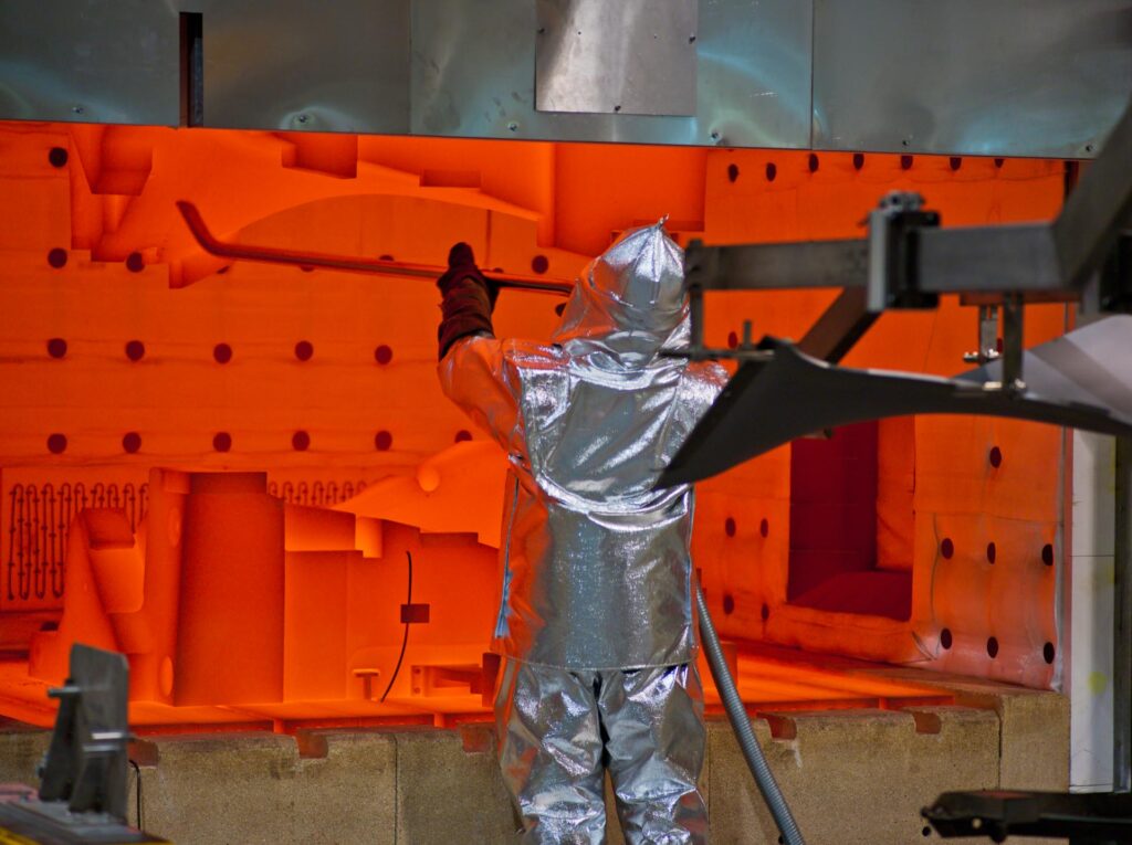 Worker in an aluminised heat suit at the open door of a glowing orange furnace during fan blade production at the Rolls-Royce Seletar fan blade plant, Singapore