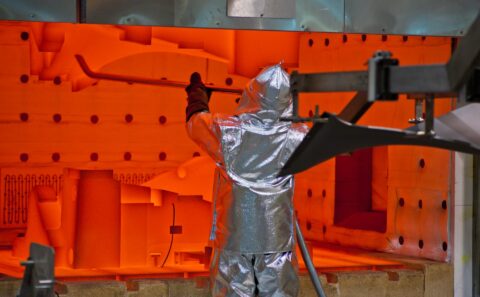 Worker in an aluminised heat suit at the open door of a glowing orange furnace during fan blade production at the Rolls-Royce Seletar fan blade plant, Singapore