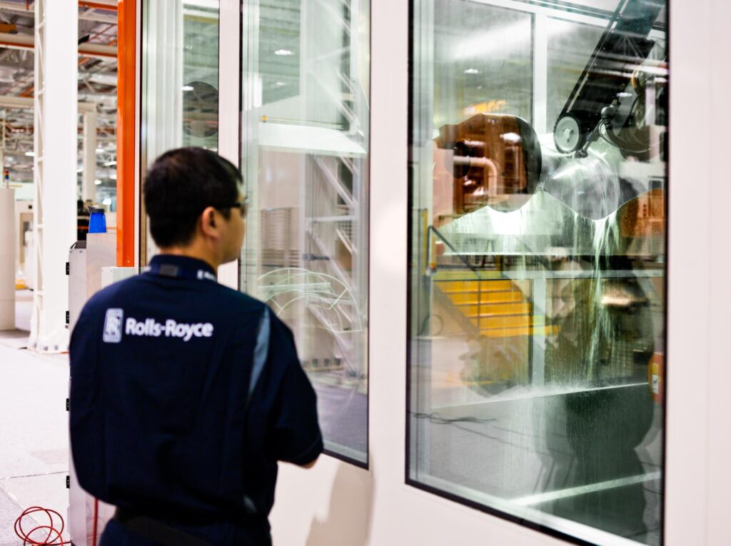 Rolls-Royce technician seen from behind, supervising the robotic water-jet polishing of a fan blade through the glass of the cell at the Seletar fan blade plant, Singapore