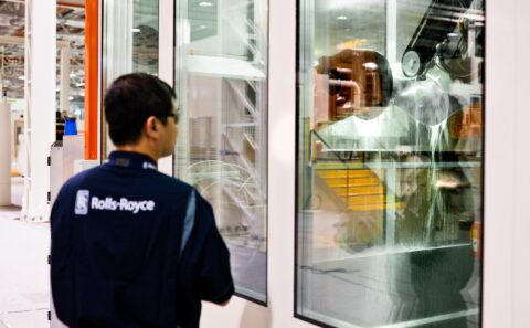 Rolls-Royce technician seen from behind, supervising the robotic water-jet polishing of a fan blade through the glass of the cell at the Seletar fan blade plant, Singapore